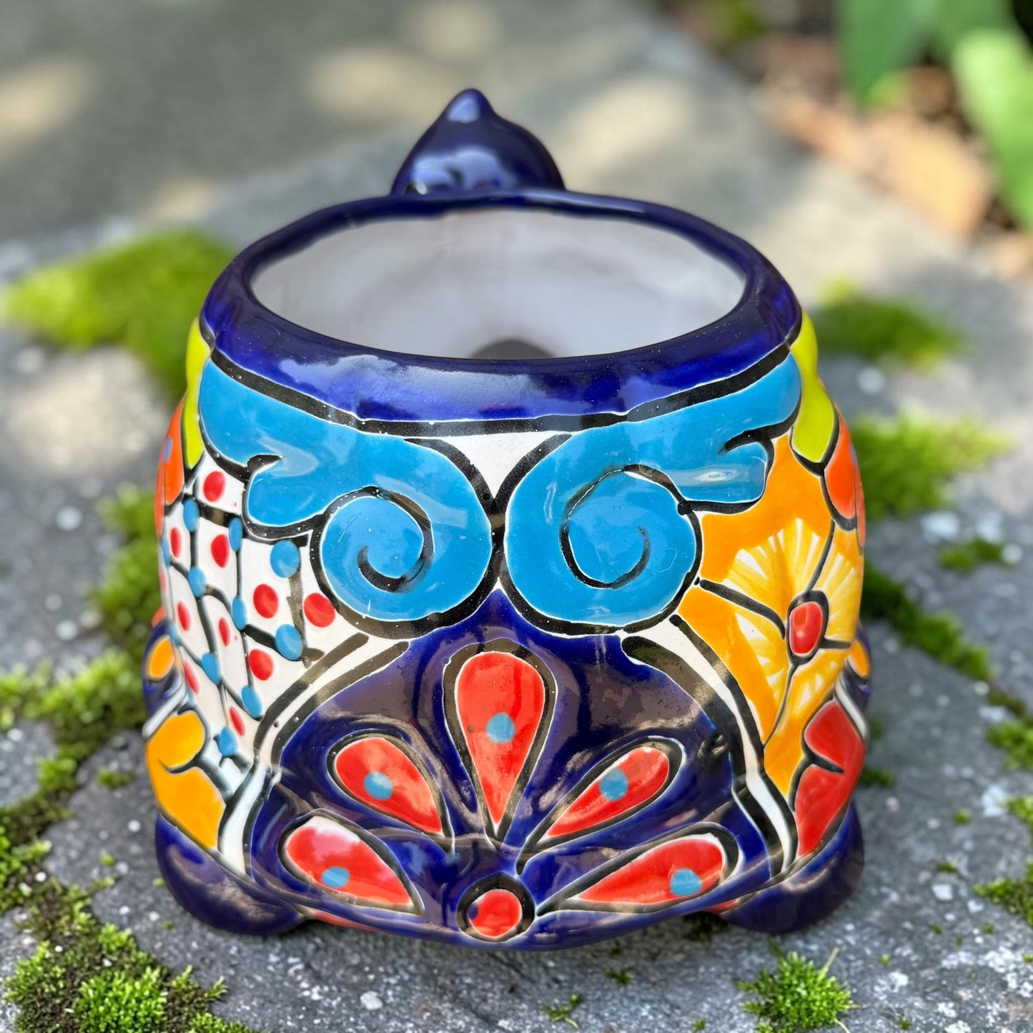 A colorful talavera turtle planter with a hand-painted Mexican-style pattern in orange, blue, yellow, and white, sitting on a mossy stone outdoors. Back of turtle shell.