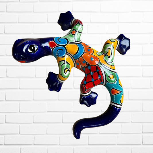 A colorful ceramic wall sculpture of a stylized lizard, decorated with vibrant hand-painted patterns in blue, orange, green, yellow, and red, mounted on a white brick wall.