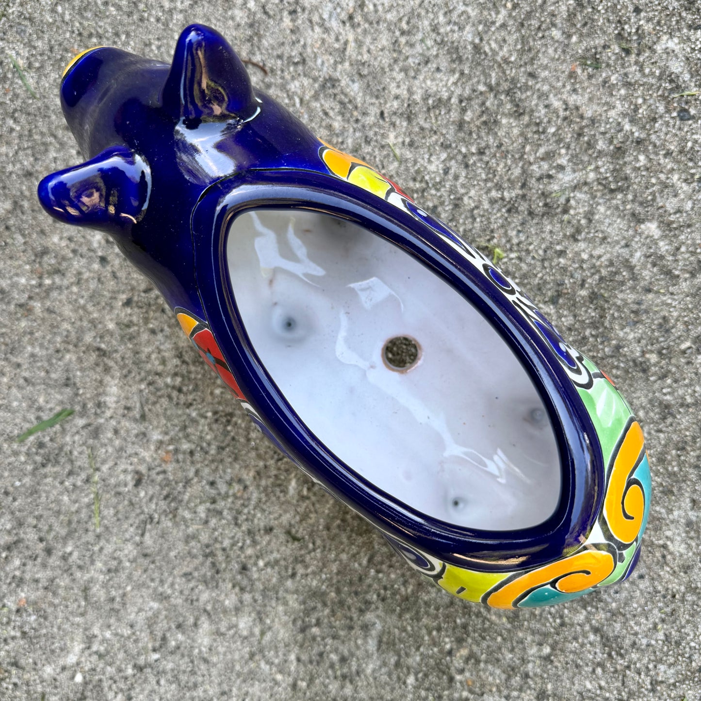 A glossy talavera pig figurine decorated with vibrant hand-painted patterns in blue, yellow, red, green, and white, standing on a brick surface with a blurred outdoor garden background, top opening of pot.