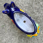 A glossy talavera pig figurine decorated with vibrant hand-painted patterns in blue, yellow, red, green, and white, standing on a brick surface with a blurred outdoor garden background, top opening of pot.