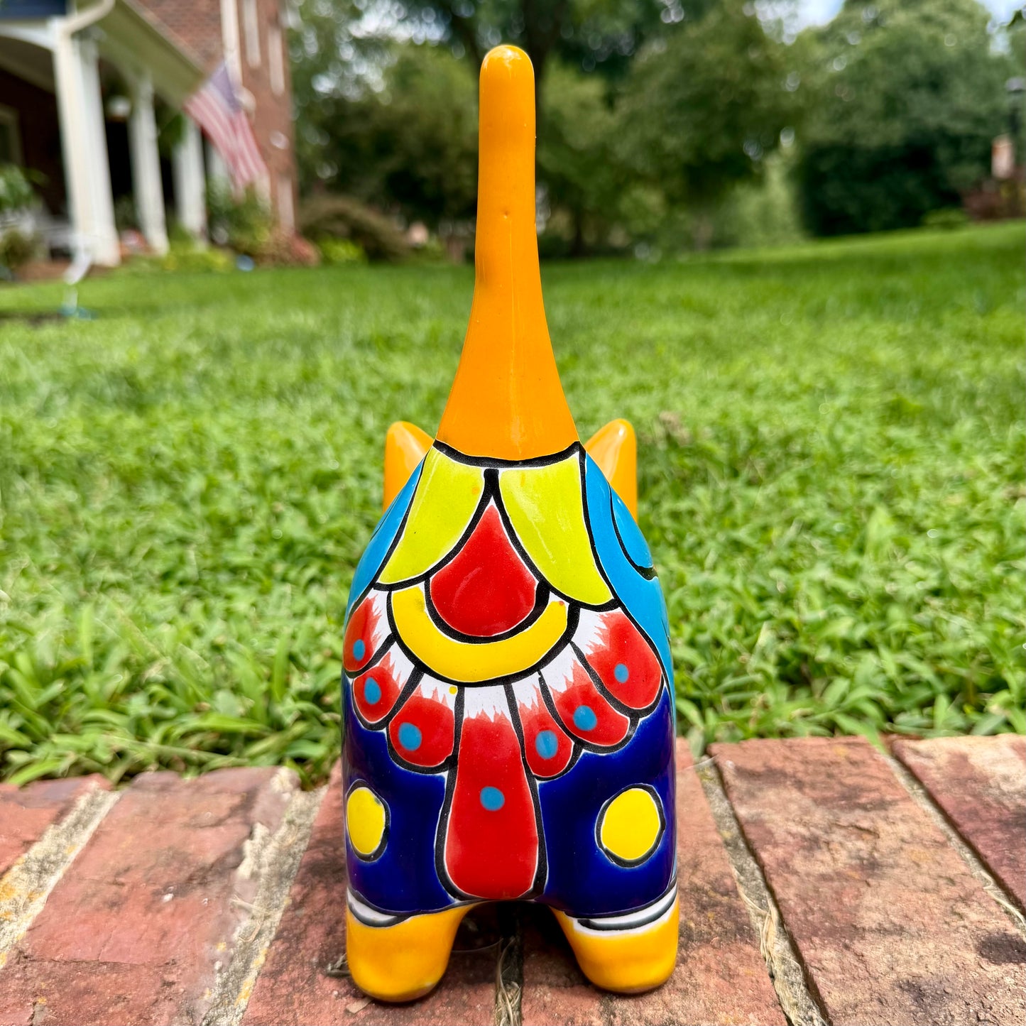 A hand-painted ceramic cat figurine in bright yellow with red, blue, and beige patterns, standing on a brick surface with a blurred green lawn and house in the background, cocktail view of cat.