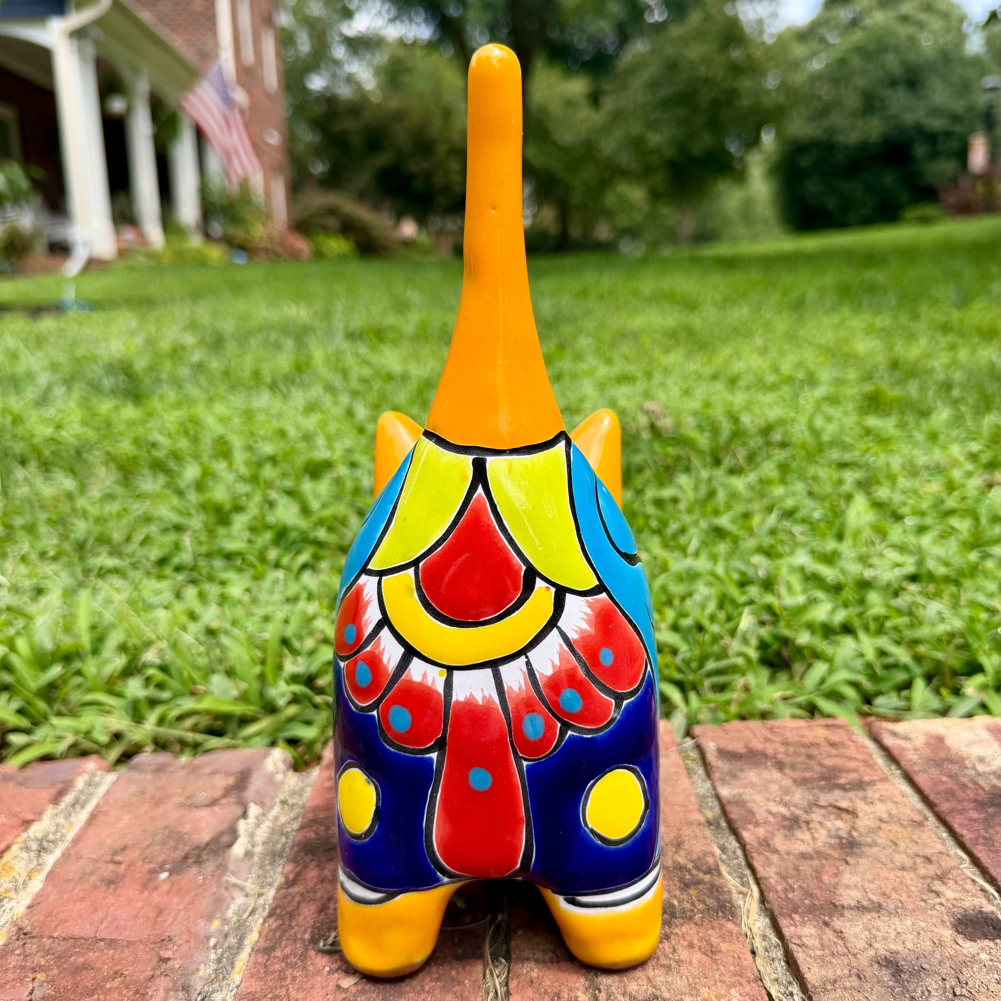 A hand-painted ceramic cat figurine in bright yellow with red, blue, and beige patterns, standing on a brick surface with a blurred green lawn and house in the background, cocktail view of cat.