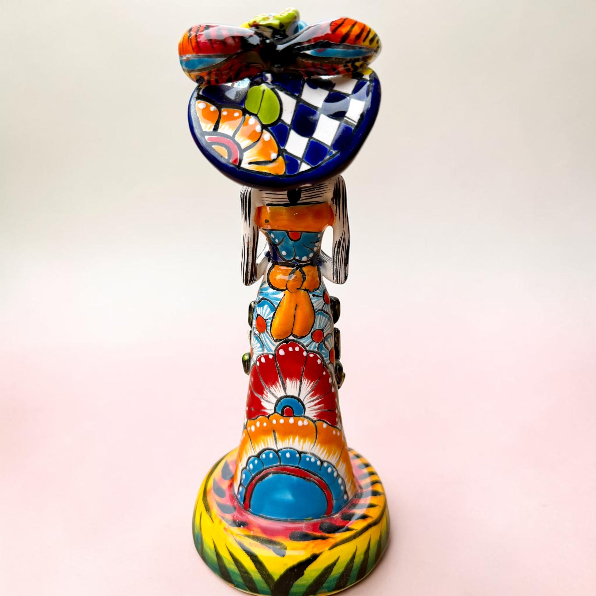 A Talavera Day of the Dead (Día de los Muertos) ceramic skeleton figurine with calla Lillies standing on a patterned base against a soft pink gradient background. This is her back view showing a bow at her waist.