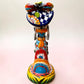 A Talavera Day of the Dead (Día de los Muertos) ceramic skeleton figurine with calla Lillies standing on a patterned base against a soft pink gradient background. This is her back view showing a bow at her waist.
