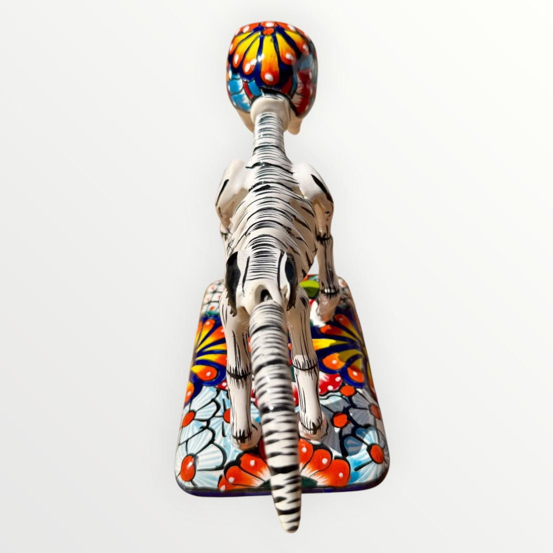 A ceramic Mexican-style skeleton dog figurine standing on a colorful floral base against a plain white background. Back view of catrina dog showing backbone.