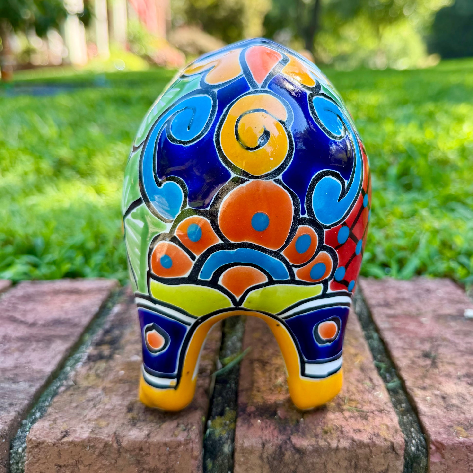 A colorful ceramic pig planter with a yellow-orange face and body, decorated with blue, green, red, and white floral patterns, sitting on a brick surface with a blurred outdoor garden background. Photo of the pigs back tail. Left side of pig pot.