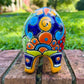 A colorful ceramic pig planter with a yellow-orange face and body, decorated with blue, green, red, and white floral patterns, sitting on a brick surface with a blurred outdoor garden background. Photo of the pigs back tail. Left side of pig pot.