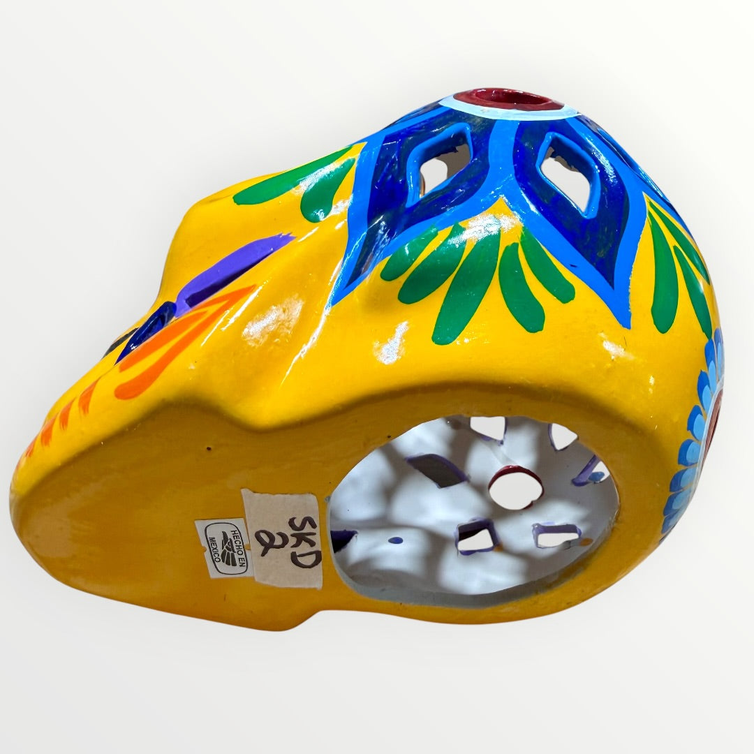 A hand-painted ceramic sugar skull in vibrant yellow, decorated with colorful floral and geometric patterns in blue, purple, green, orange, and red, set against a plain white background bottom of skull.