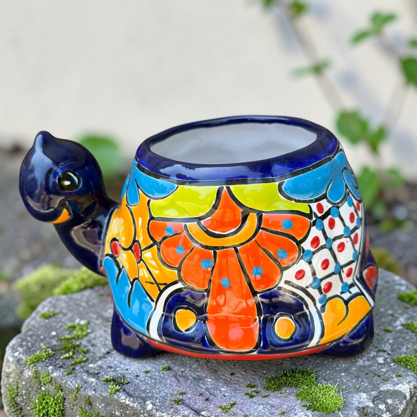 A colorful talavera turtle planter with a hand-painted Mexican-style pattern in orange, blue, yellow, and white, sitting on a mossy stone outdoors.