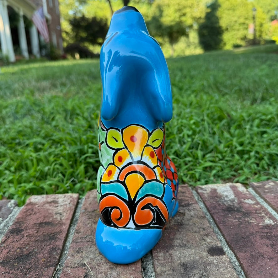 A hand-painted talavera ceramic dog figurine in a sitting position, featuring a vibrant blue base with colorful geometric patterns in red, yellow, orange, and white. The figurine is photographed outdoors on a brick surface with a blurred green lawn and trees in the background back view.