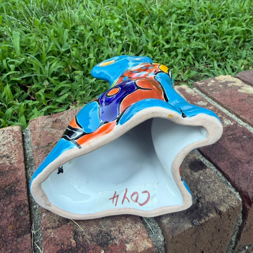 A hand-painted talavera ceramic dog figurine in a sitting position, featuring a vibrant blue base with colorful geometric patterns in red, yellow, orange, and white. The figurine is photographed outdoors on a brick surface with a blurred green lawn and trees in the background bottom view