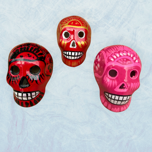 Three hand-painted Mexican sugar skulls (calaveras) in red, orange, and pink, decorated with intricate black and white patterns, set against a light blue textured background.