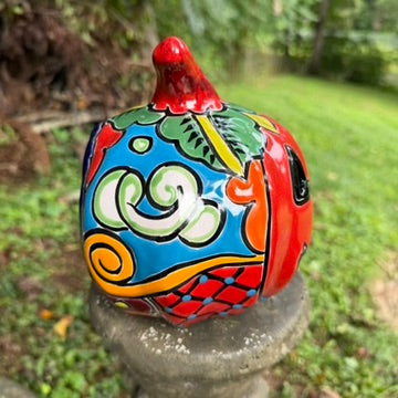 A hand-painted talavera halloween  jack-o'-lantern candle holder in red, green, black, and orange, displayed on a stone pedestal in an outdoor garden setting, left side view.