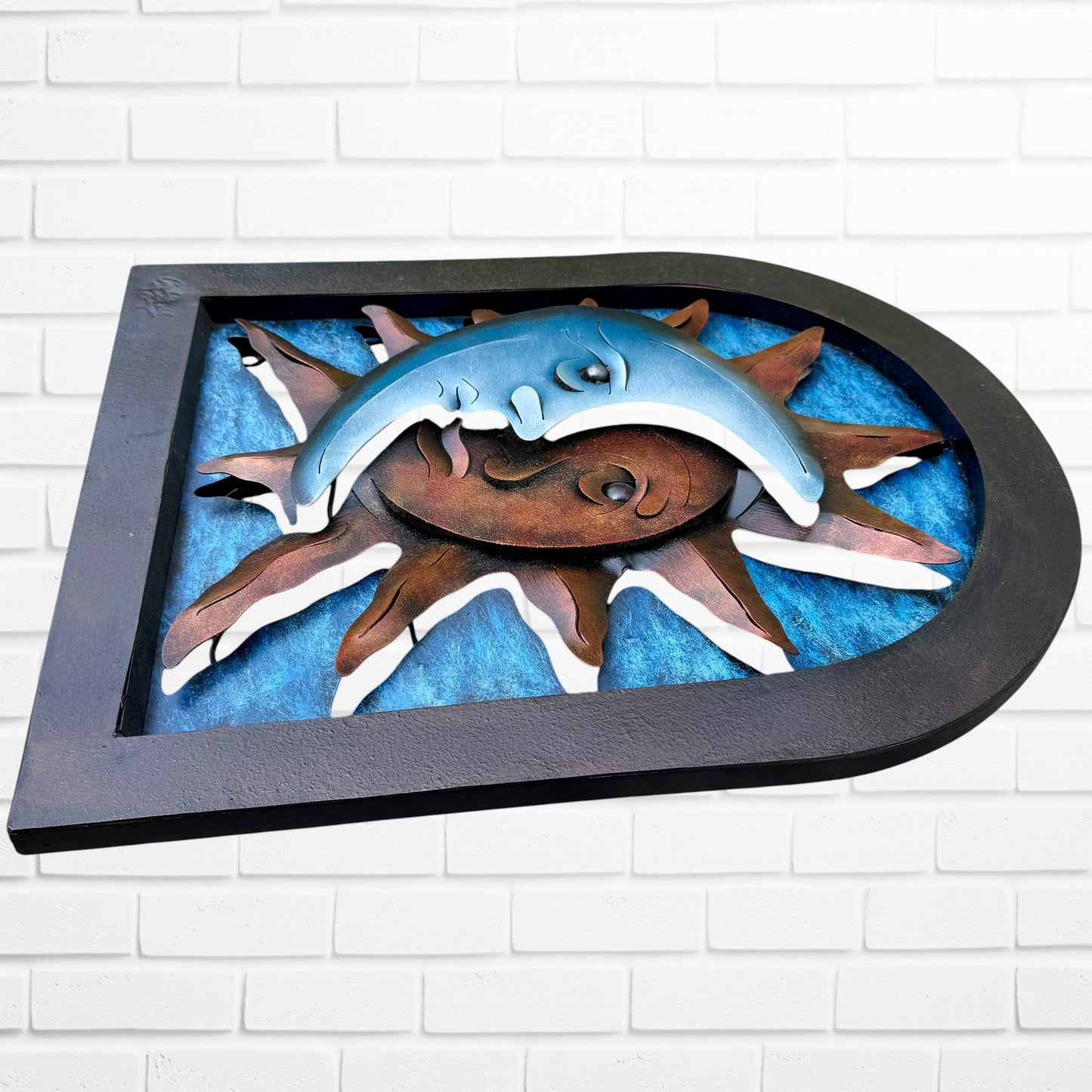 Decorative wall art of a sun and moon face with a metal frame on a white brick wall. Designed by artist Andres Martin right side view