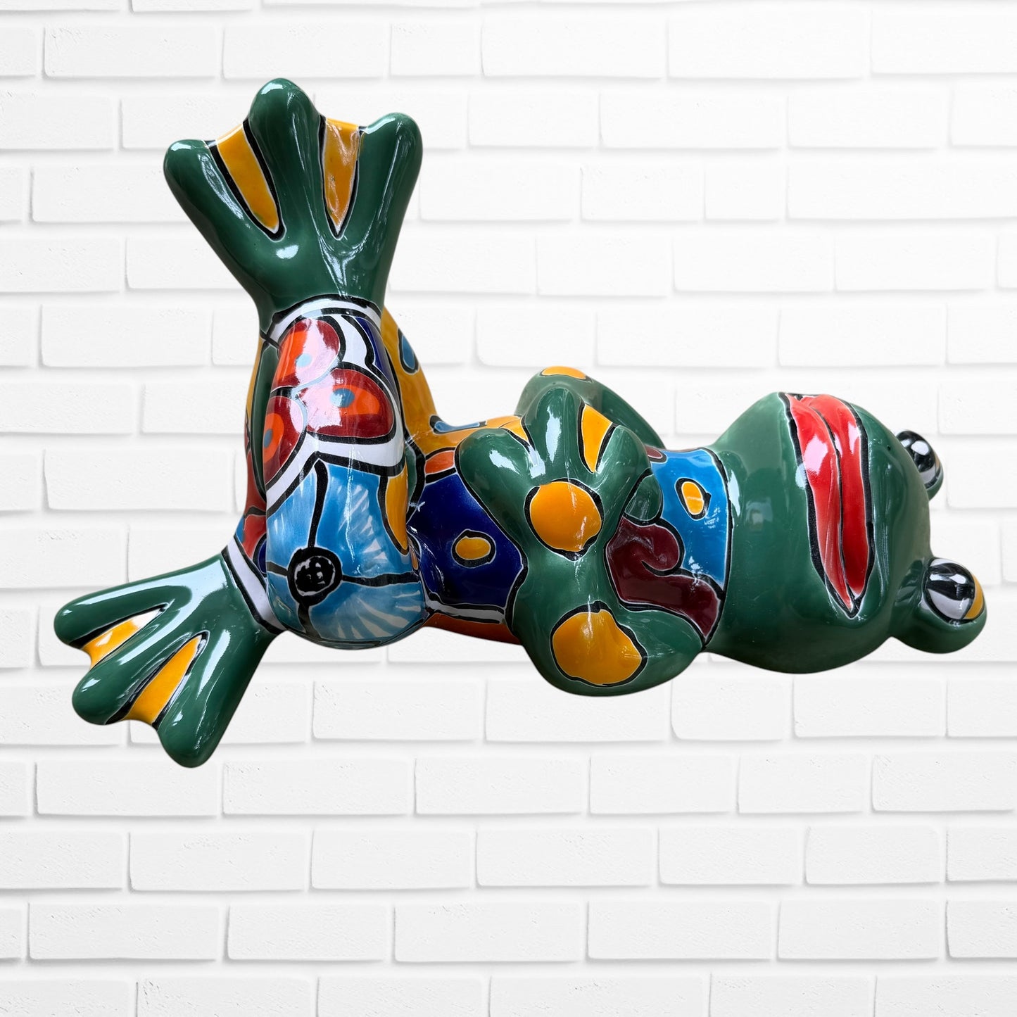 A colorful ceramic frog wall sculpture sitting cross-legged against a white painted brick wall. The frog features a glossy green body with yellow, blue, red, and left side view