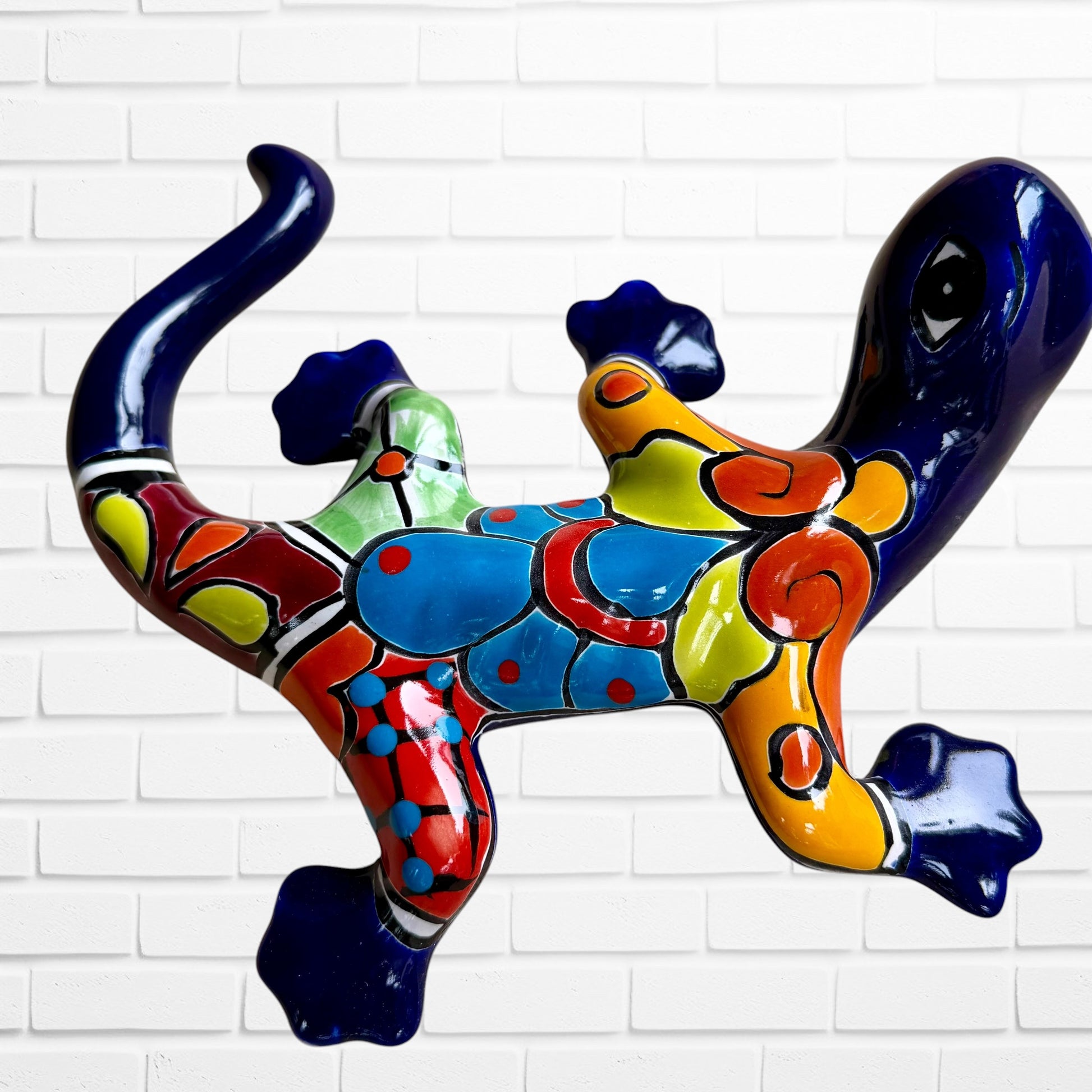 A colorful, glossy lizard wall sculpture with 
a vibrant mosaic pattern of blue, orange, yellow, green, red, and black, mounted on a white brick wall back view.