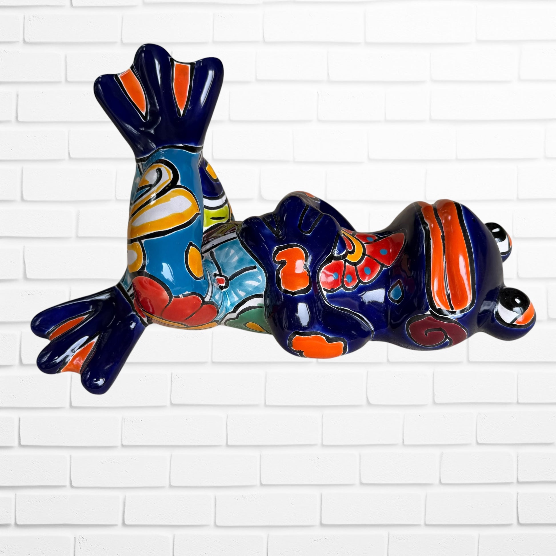 A colorful ceramic frog sculpture sitting cross-legged against a white brick wall background. The frog features a glossy deep blue body with vibrant orange, red, yellow, and light blue painted patterns and details left side view.