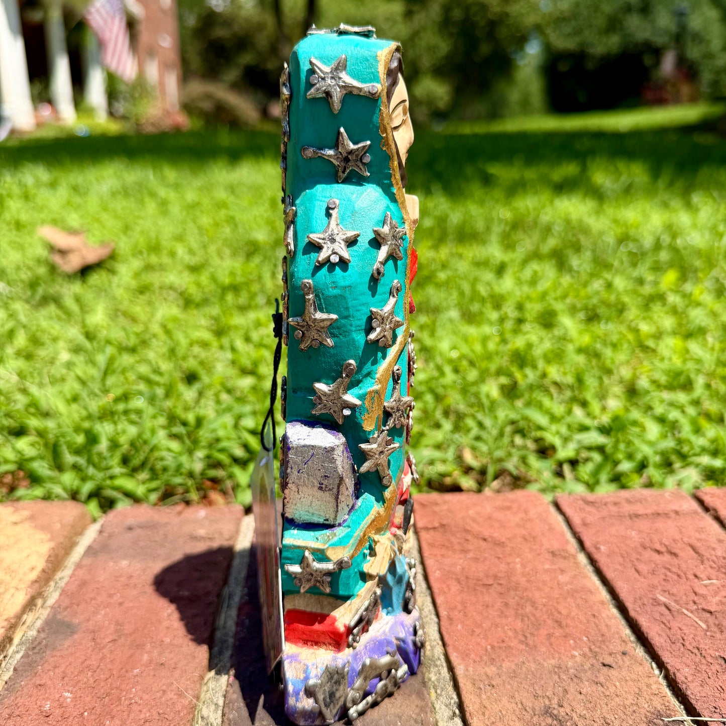 A colorful Milagros Guadalupe  (Virgin Mary) standing on a brick surface outdoors with a grassy yard in the background, left side view.