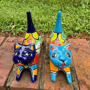 Two colorful hand-painted ceramic cats sitting on a brick ledge with a green lawn and trees in the background. One is cobalt  blue and the other is turquoise, both have traditional multi-color talavera designs, top view.