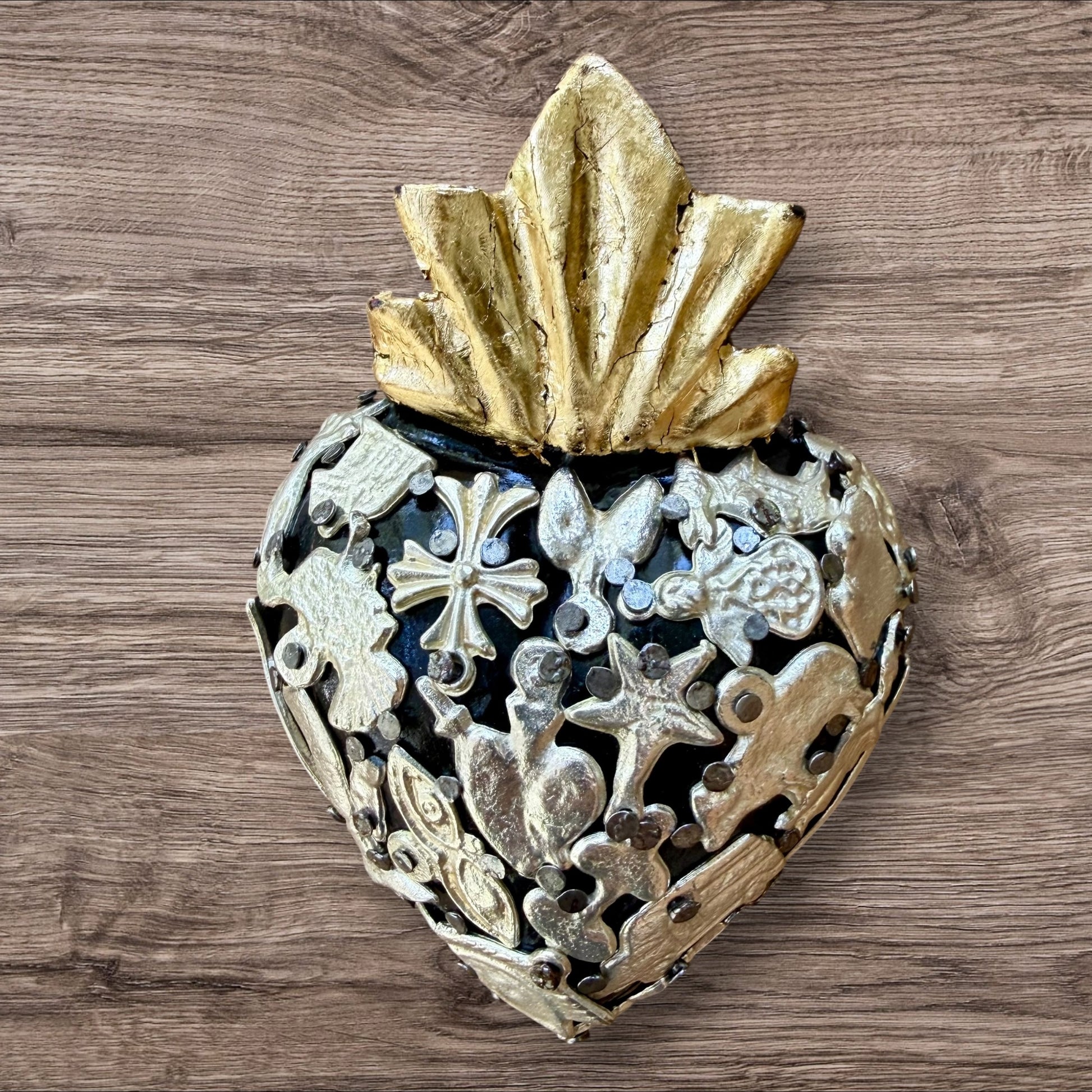 A Mexican Milagros heart in black with a gold flaming top, covered in silver charms. 