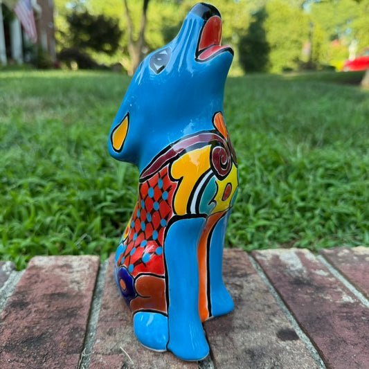 A hand-painted talavera ceramic dog figurine in a sitting position, featuring a vibrant blue base with colorful geometric patterns in red, yellow, orange, and white. The figurine is photographed outdoors on a brick surface with a blurred green lawn and trees in the background 