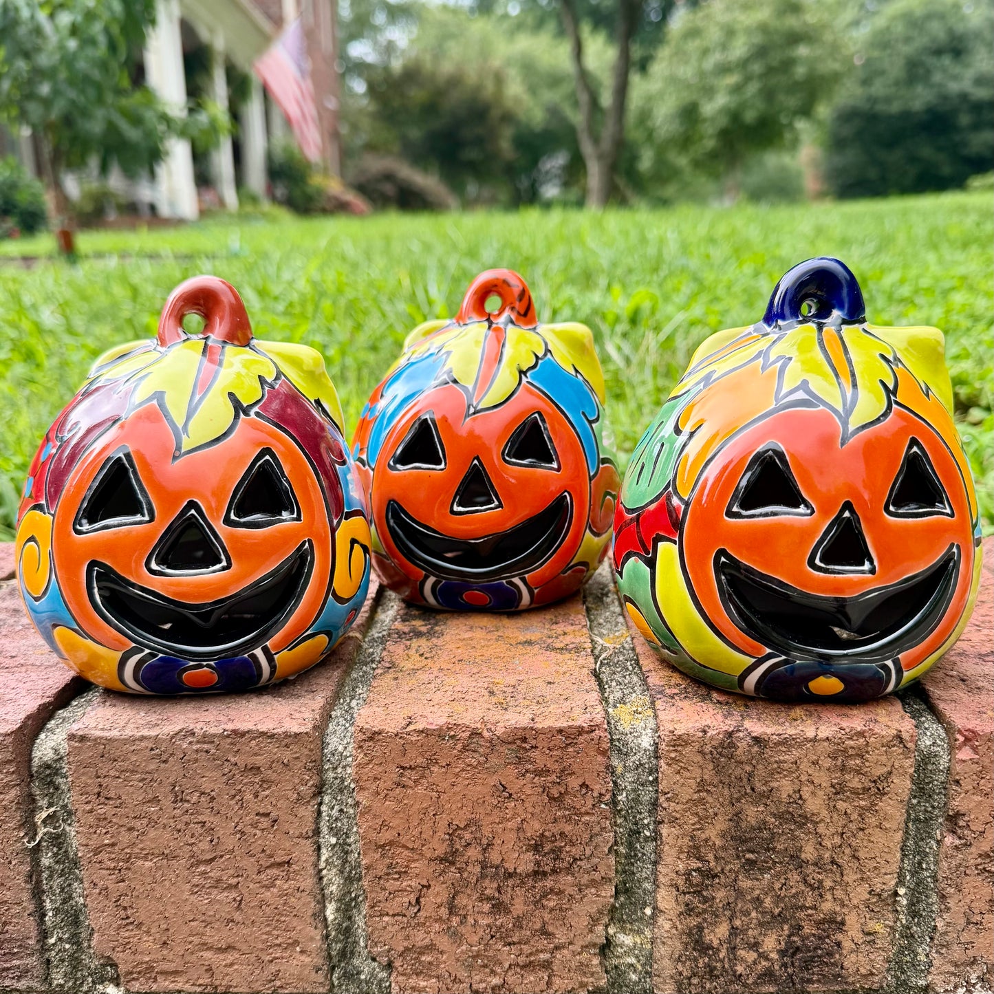 Three hand-painted Talavera ceramic jack-o'-lantern pumpkins with orange faces and yellow, blue, and red green designs, sitting on a brick path in a grassy backyard.