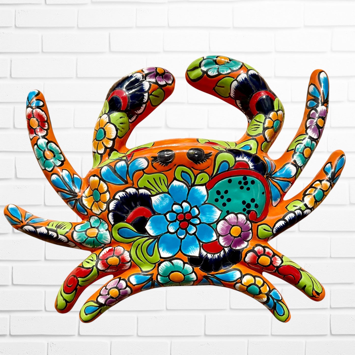 A colorful multicolor hand-painted wall art sculpture of a crab, decorated with vibrant floral patterns, shown against a white brick wall background, top shell.