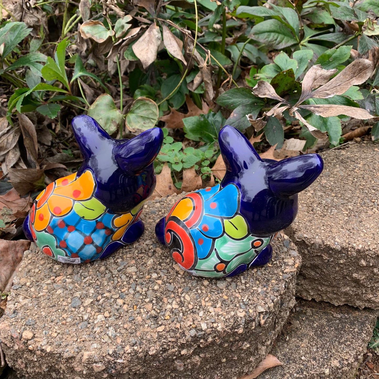 Two colorful Mexican Talavera rabbit figurines on a stone surface with greenery in the background in azul back view