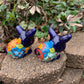 Two colorful Mexican Talavera rabbit figurines on a stone surface with greenery in the background in azul back view