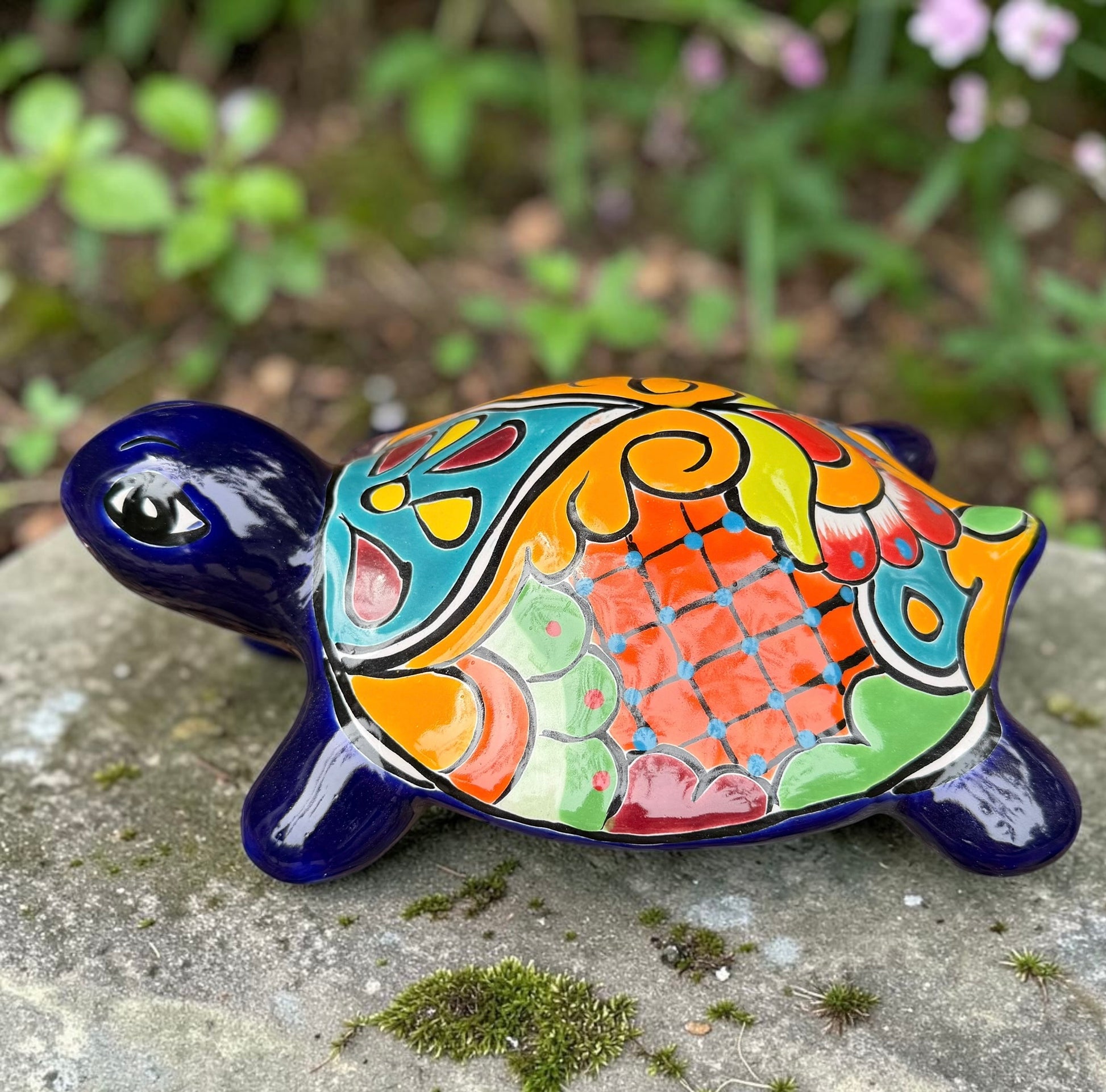 A glossy ceramic turtle figurine with a vibrant hand-painted patterned shell and a deep blue head and legs, resting on a gray stone surface with a blurred green garden background, right side view.