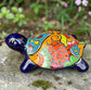 A glossy ceramic turtle figurine with a vibrant hand-painted patterned shell and a deep blue head and legs, resting on a gray stone surface with a blurred green garden background, right side view.