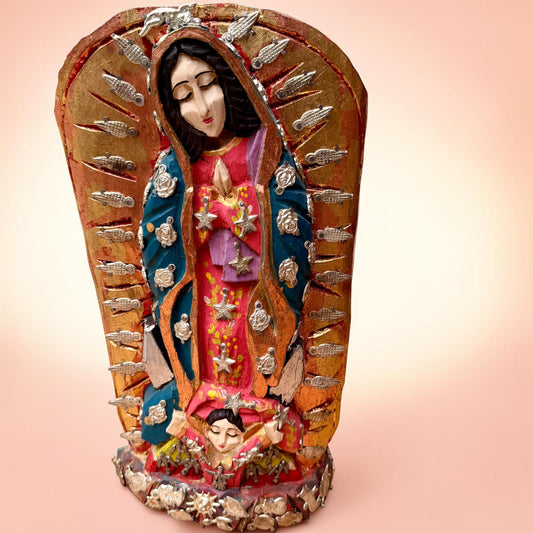 A wood religious milagros figurine of  Our Lady of Guadalupe  surrounded by a halo with silver charm embellishments, set against a soft peach gradient background.