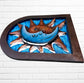 Decorative wall art of a sun and moon face with a metal frame on a white brick wall. Designed by artist Andres Martin left side view