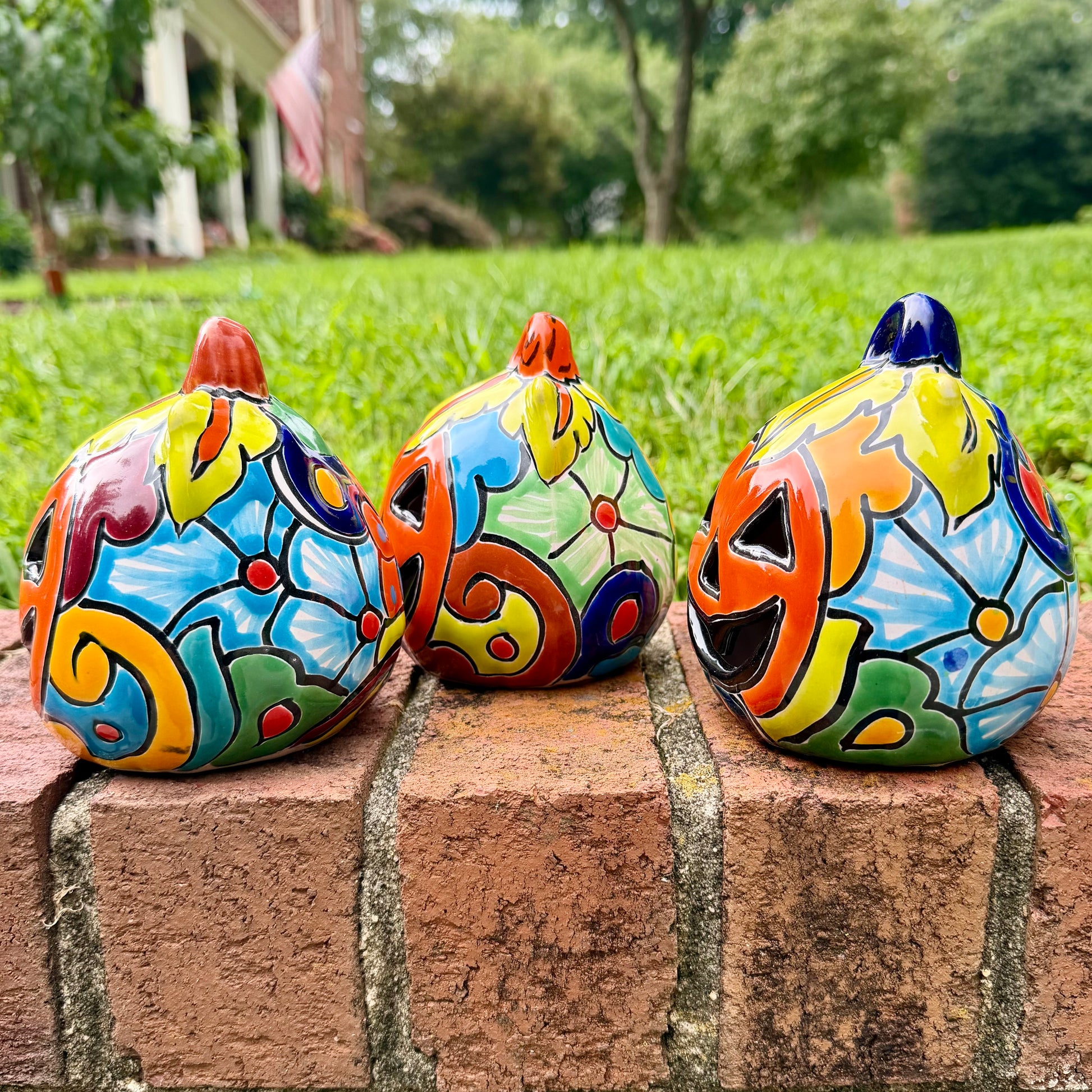 Three hand-painted Talavera ceramic jack-o'-lantern pumpkins with orange faces and yellow, blue, and red green designs, sitting on a brick path in a grassy backyard, right side view.