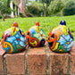 Three hand-painted Talavera ceramic jack-o'-lantern pumpkins with orange faces and yellow, blue, and red green designs, sitting on a brick path in a grassy backyard, right side view.
