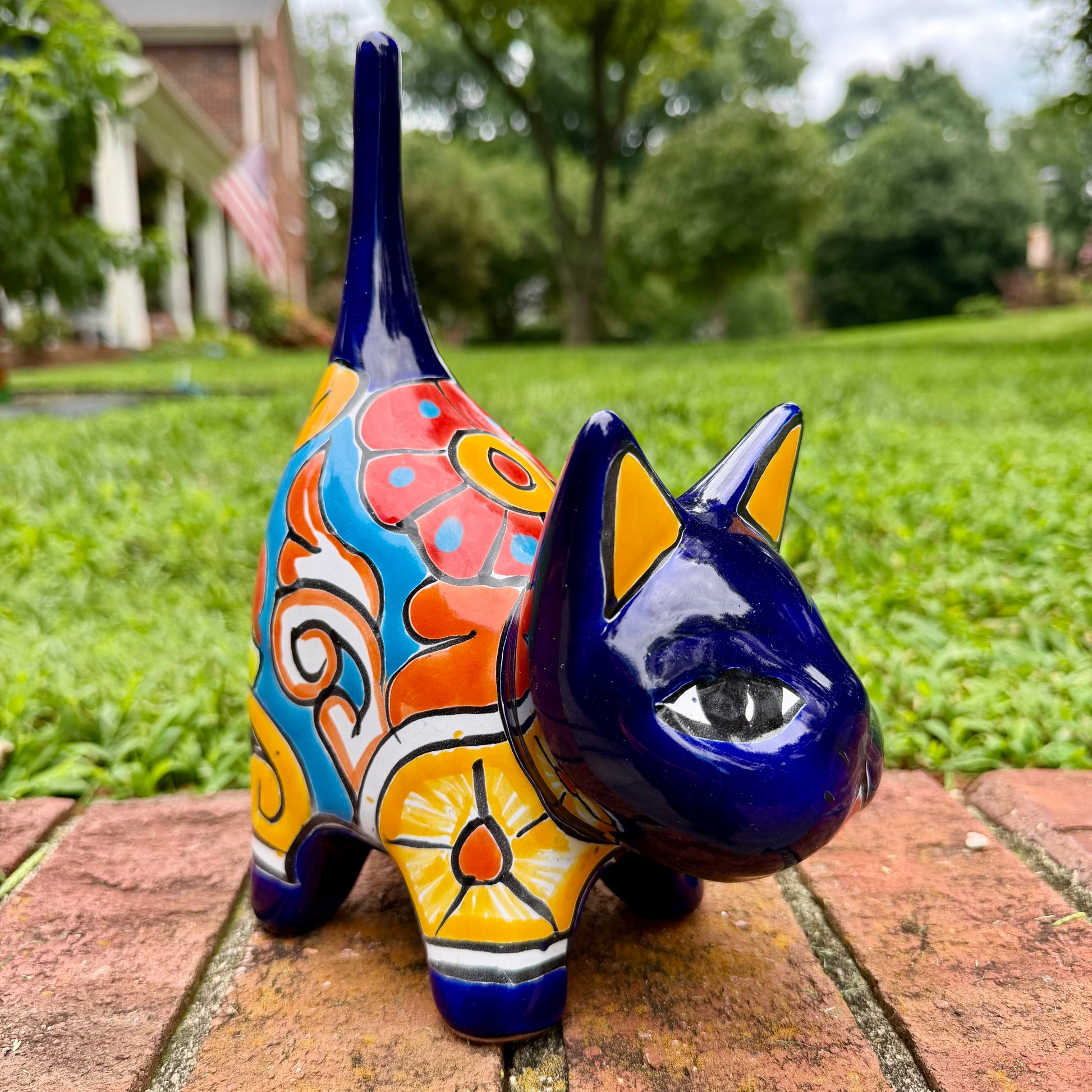 A glossy talavera ceramic cat figurine decorated with vibrant hand-painted patterns in blue, orange, yellow, and red, standing on a brick path with a green lawn and a house in the blurred background.