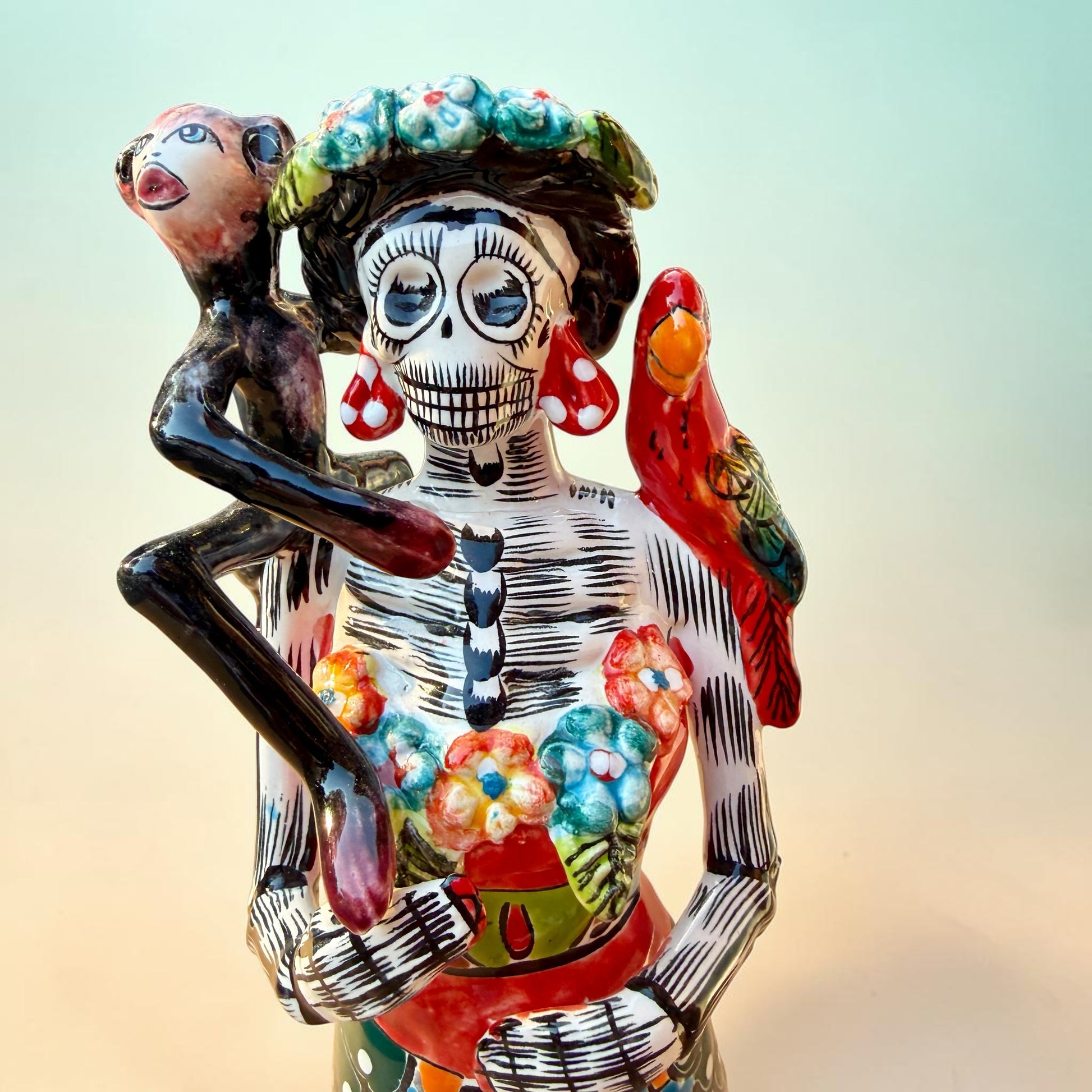 A Talavera Day of the Dead (Día de los Muertos) Frida figurine  with a monkey on her shoulder and a red parrot on her other shoulder, set against a light blue gradient background. Close up of Frida’s face.