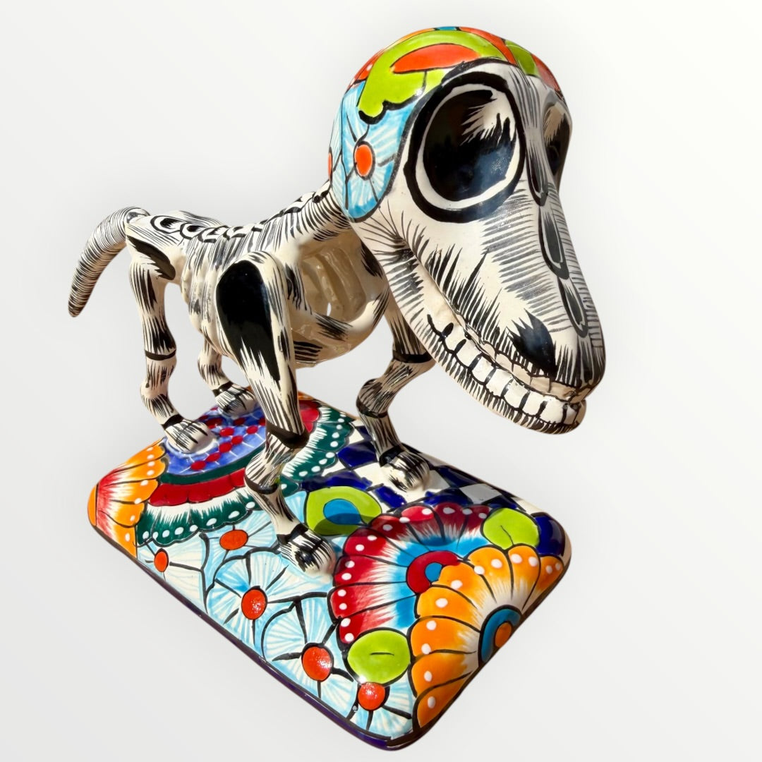 A talavera catrina dog skeleton figurine with colorful hand-painted floral and geometric patterns, standing on a matching decorated base against a plain white background.