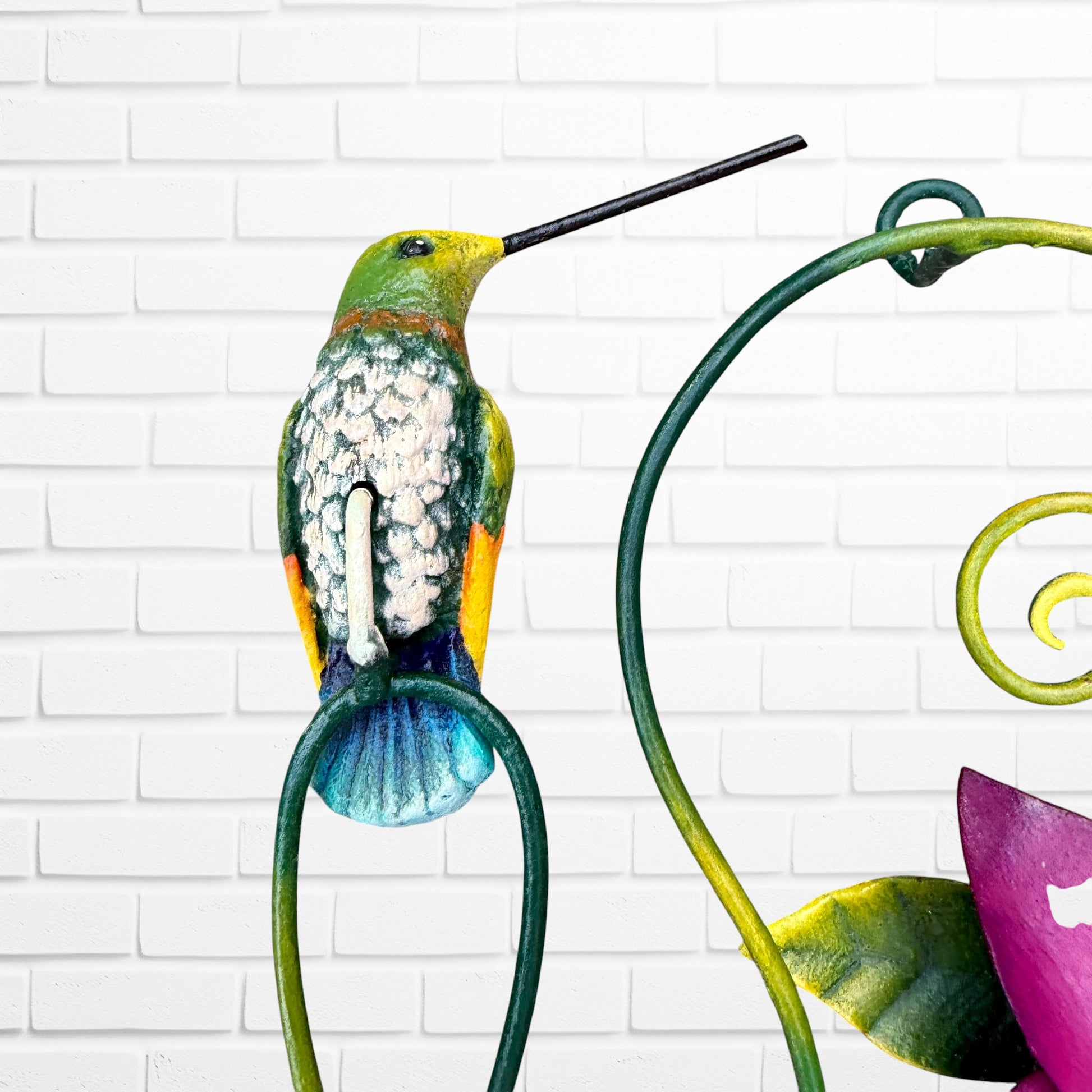 Decorative wall art with purple flowers and colorful hummingbirds on a white brick background top bird close up