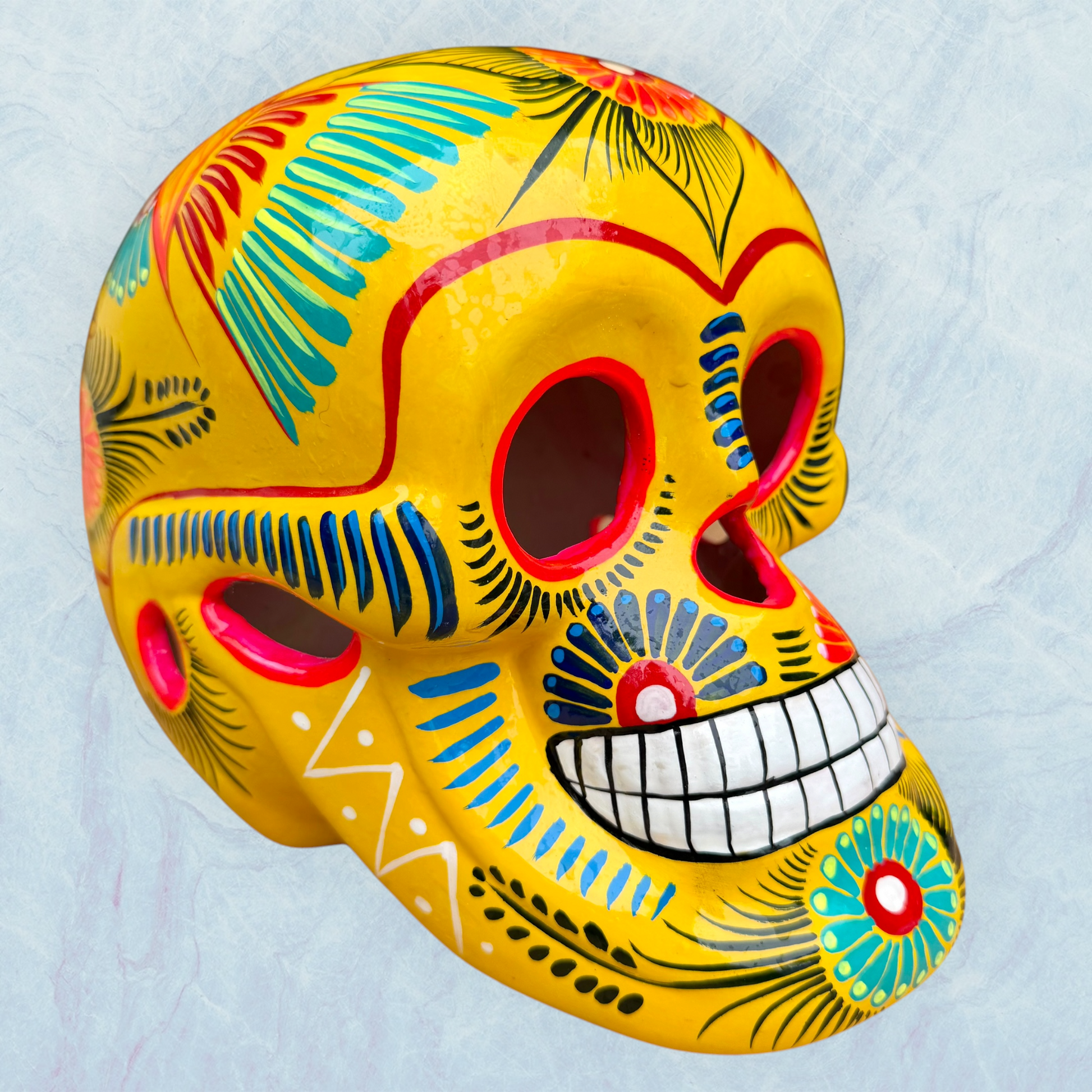 A vibrant yellow sugar skull decorated with colorful floral and geometric patterns in blue, red, black, and white, set against a light blue textured background.