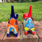 Two colorful ceramic cat figurines, one blue and one red, decorated with geometric patterns, displayed on a brick ledge with a green lawn in the background back tail view.
