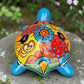 A hand-painted ceramic turtle figurine resting on a mossy stone outdoors, featuring a bright blue body and a highly decorated shell with vibrant orange, yellow, red, and blue patterns, back view of Shell.