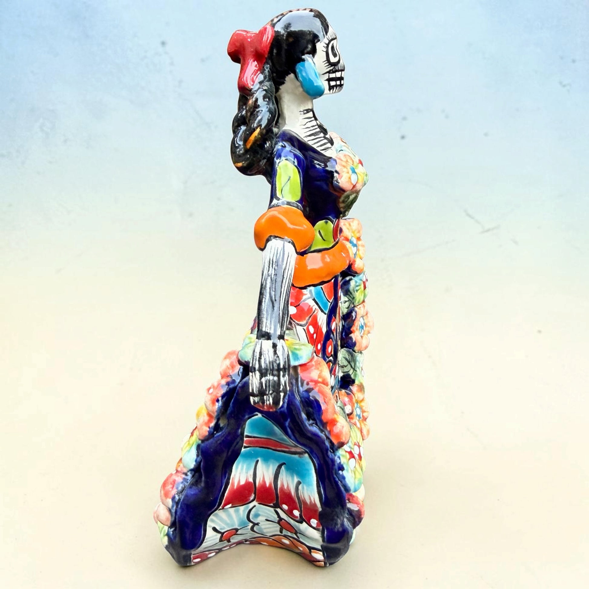 A colorful talavera Day of the Dead (Día de los Muertos) dancing skeleton ballerina figurine. The figurine features black hair in braids wearing a vibrant multicolor dress with floral patterns and orange accents, set against a light blue and beige gradient background, left side view.