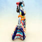 A colorful talavera Day of the Dead (Día de los Muertos) dancing skeleton ballerina figurine. The figurine features black hair in braids wearing a vibrant multicolor dress with floral patterns and orange accents, set against a light blue and beige gradient background, left side view.