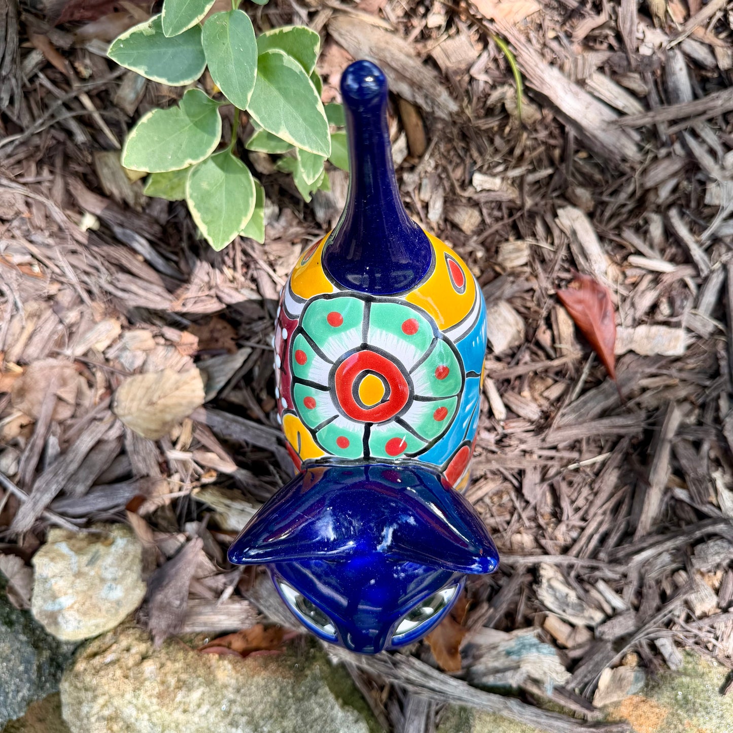 A colorful ceramic cat figurine with a glossy blue head and tail, yellow ears and paws, and a patterned body, standing outdoors on mulch with green plants in the background, top view of cat,
