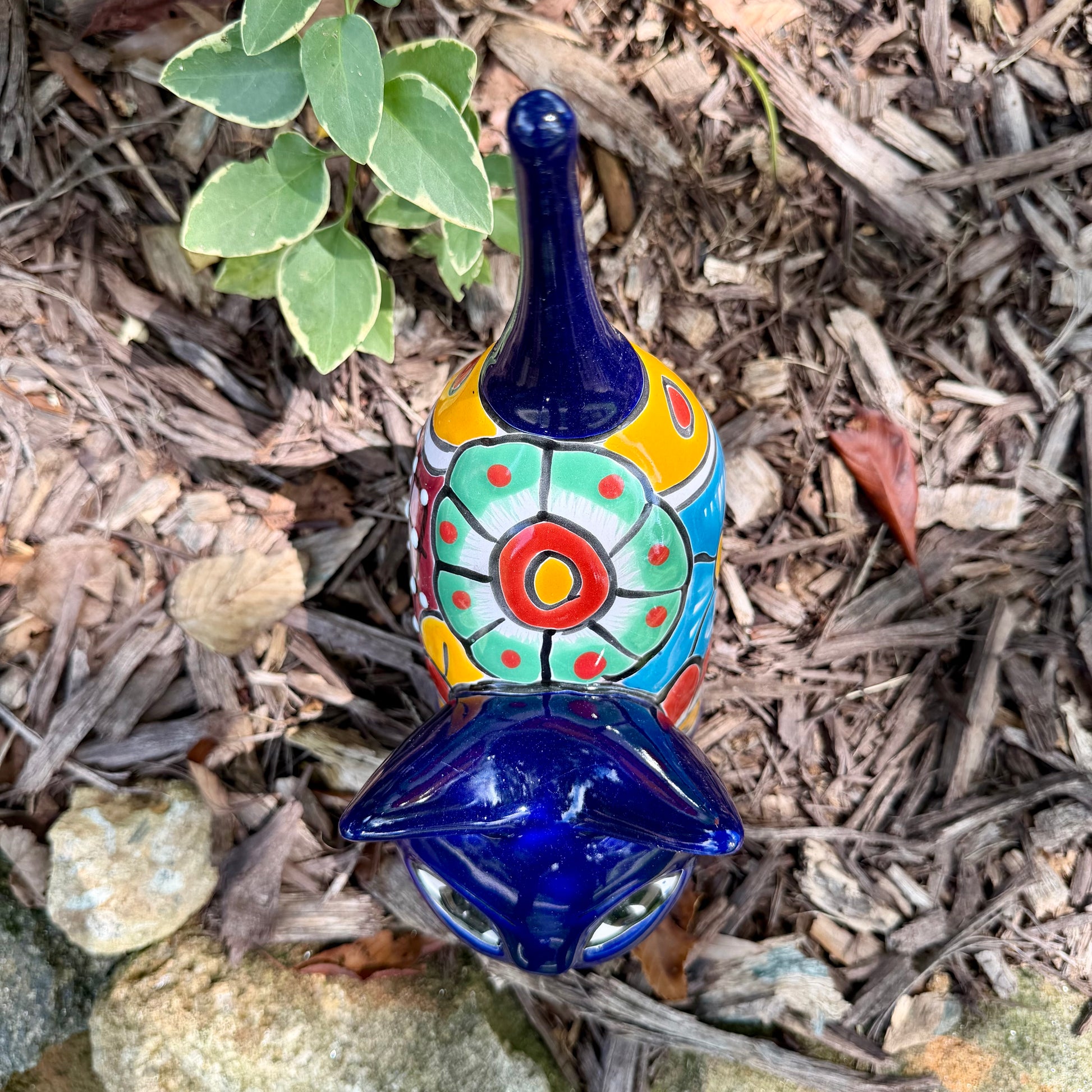 A colorful ceramic cat figurine with a glossy blue head and tail, yellow ears and paws, and a patterned body, standing outdoors on mulch with green plants in the background, top view of cat,