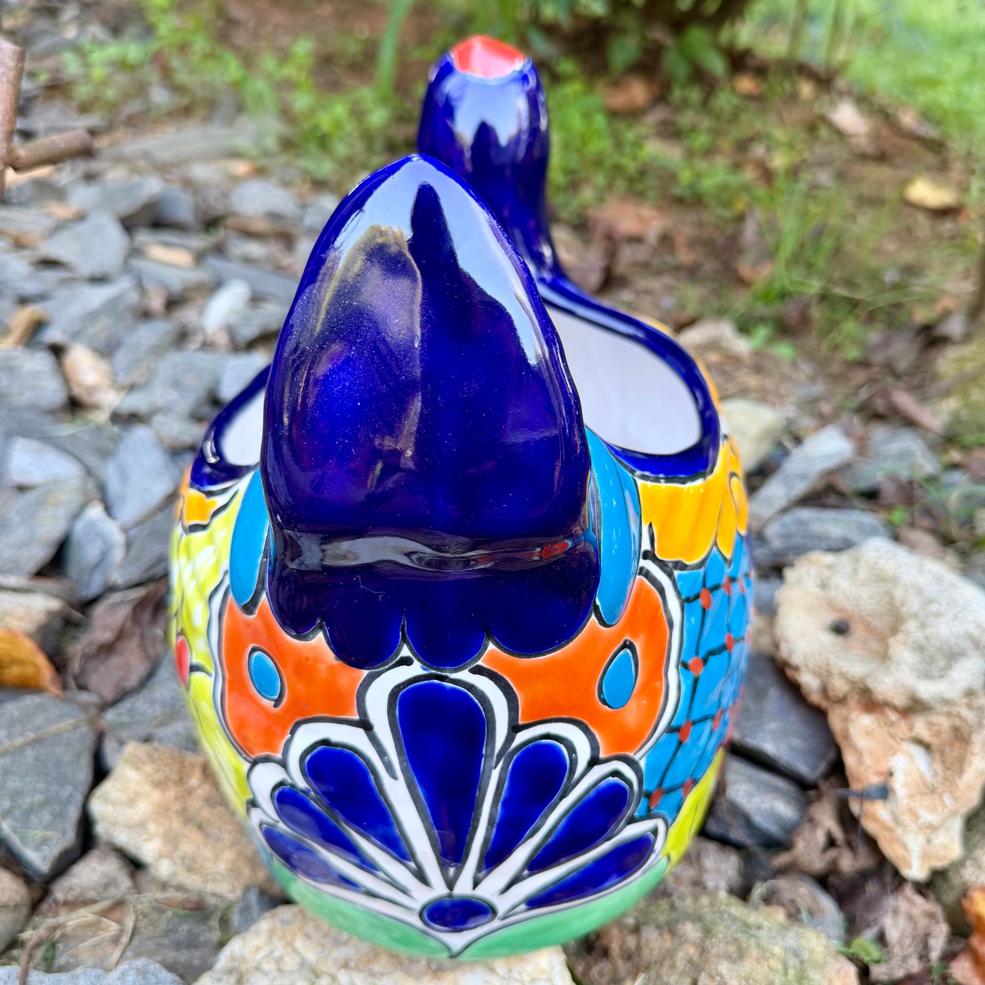 A colorful ceramic chicken-shaped planter with a glossy hand-painted finish, placed outdoors on a bed of small gray rocks and greenery, back tail view.