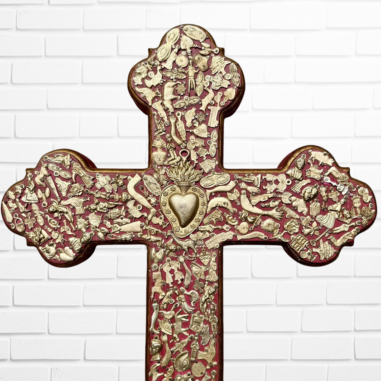 Decorative Milagros wood cross with many charms “miracles” on a white brick wall background Top half