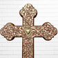 Decorative Milagros wood cross with many charms “miracles” on a white brick wall background Top half