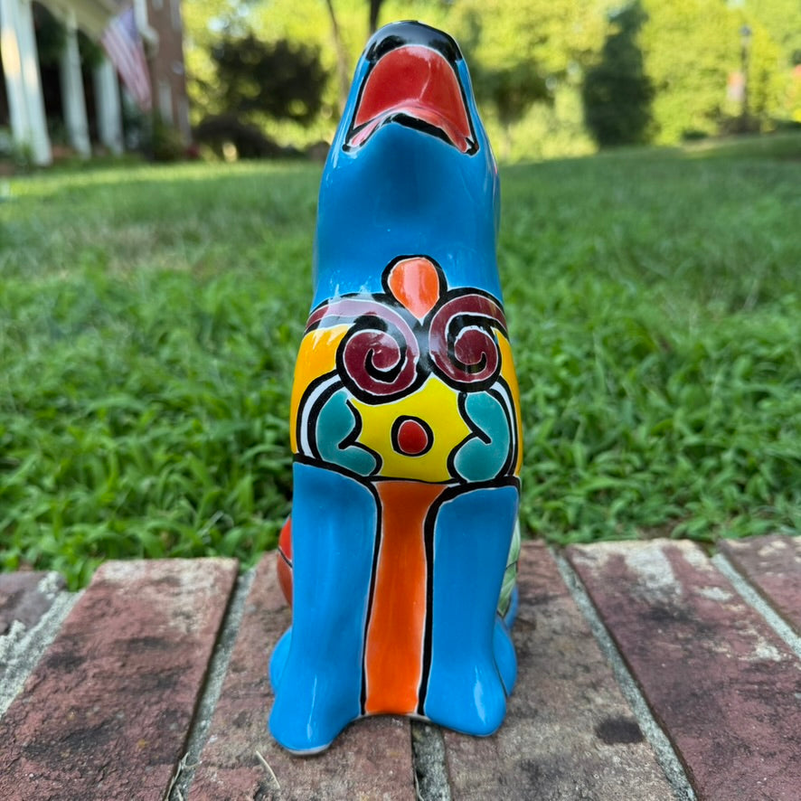 A hand-painted talavera ceramic dog figurine in a sitting position, featuring a vibrant blue base with colorful geometric patterns in red, yellow, orange, and white. The figurine is photographed outdoors on a brick surface with a blurred green lawn and trees in the background front view